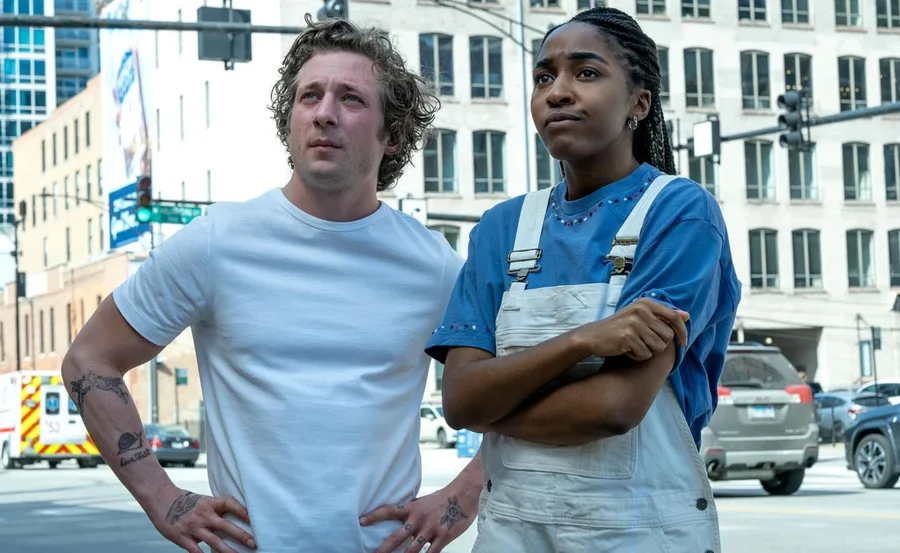 Two people standing on a city street, one in a white shirt and one in blue overalls, both looking intently ahead.