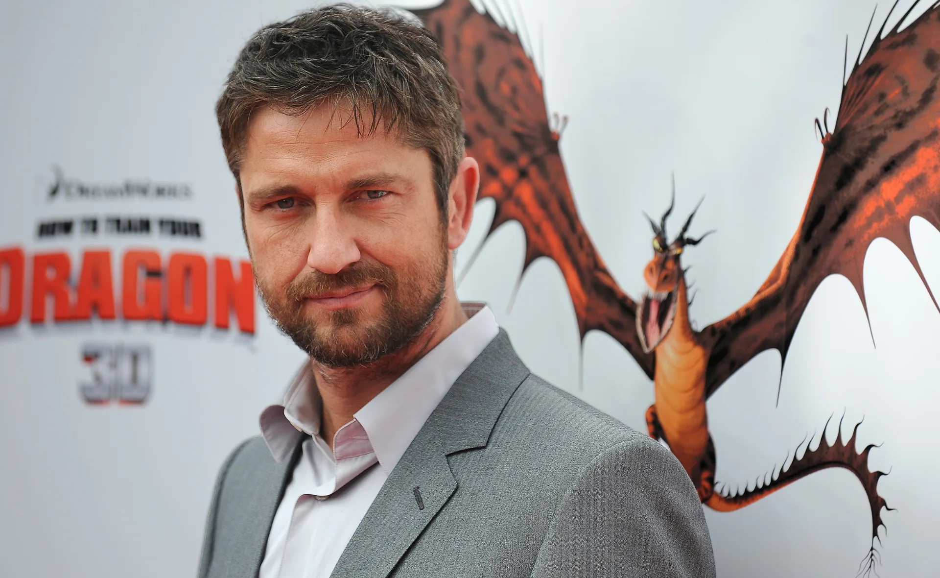 Man in gray suit at "How to Train Your Dragon" event with backdrop of flying dragon.