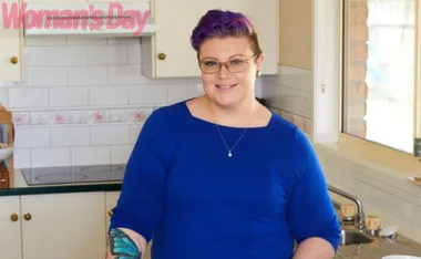 Person with purple hair, wearing glasses and blue shirt, stands in kitchen beside a window. Woman's Day logo visible.