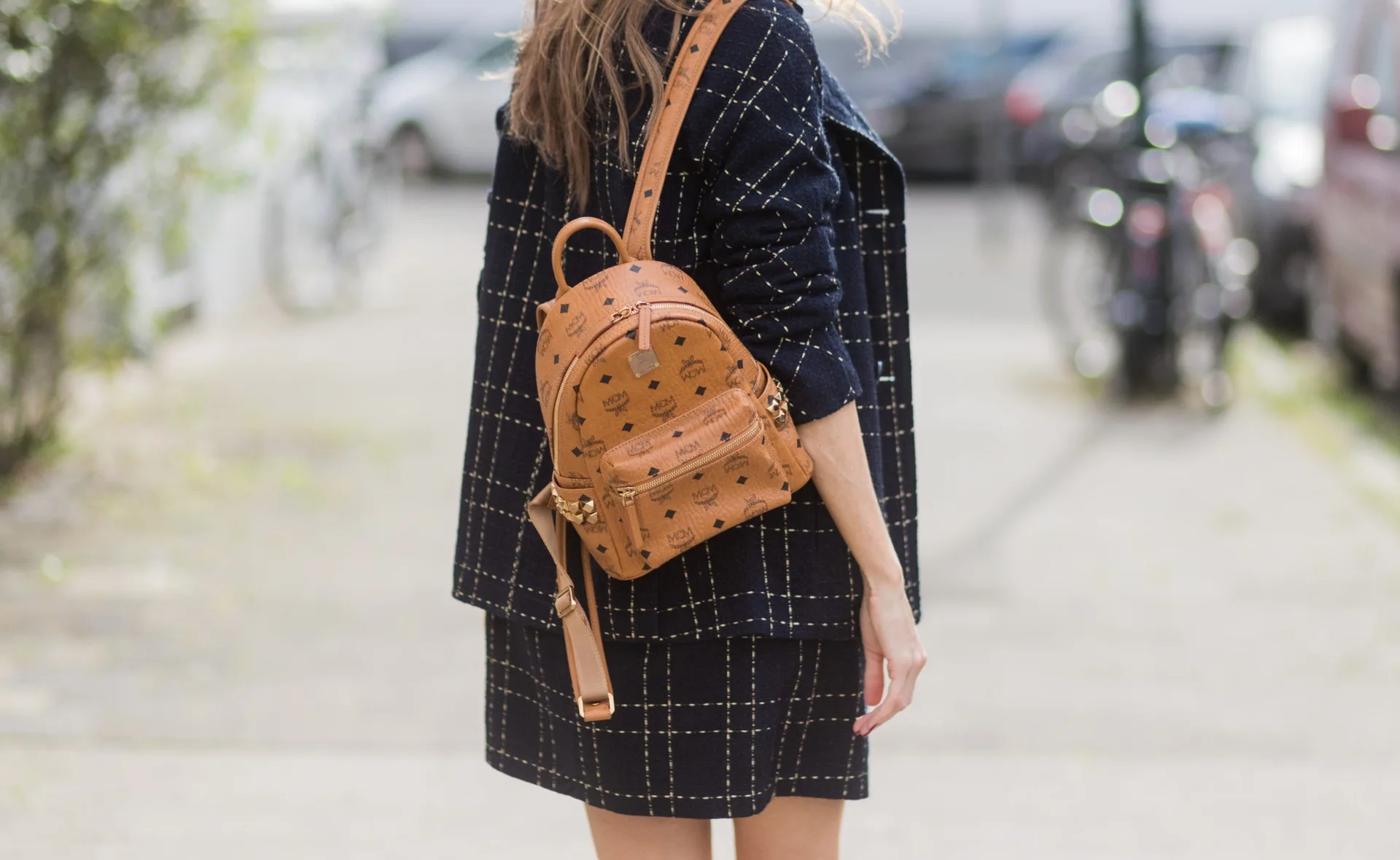 Woman in dark checkered coat wearing a brown MCM backpack, standing on a blurred urban street.