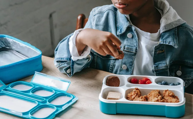 Child eating lunch from a blue bento lunchbox with various snacks and fruits on a table.