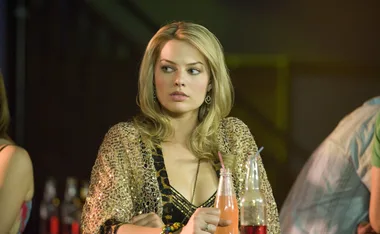 A woman with blonde hair and a sparkly top sits at a bar with drinks in front of her, looking to the side.