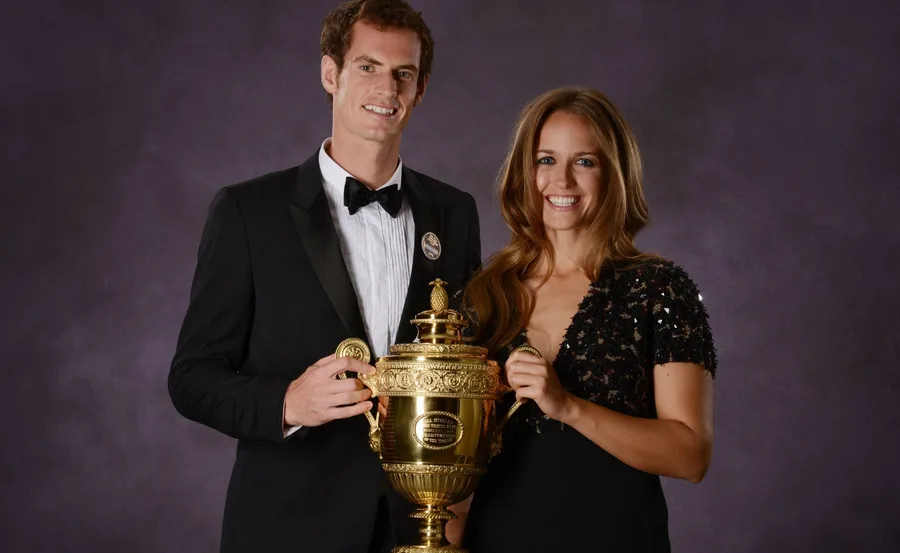 A man in a tuxedo and a woman in a black dress smile while holding a large gold trophy together.