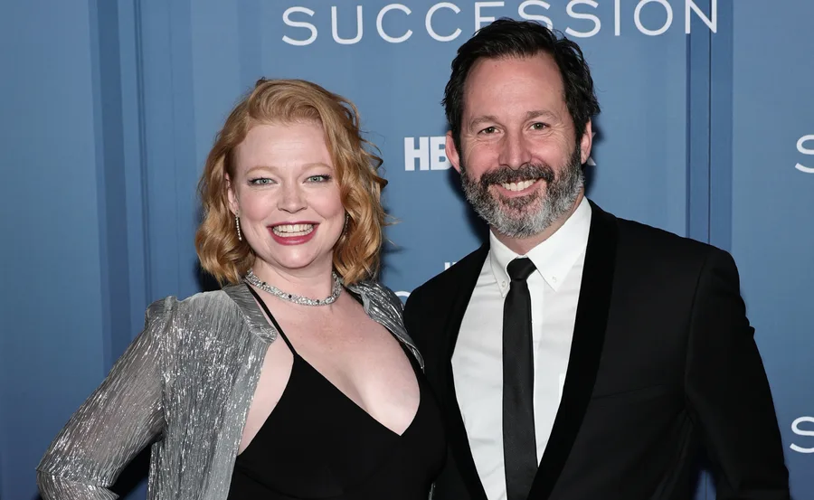 Couple smiling at "Succession" premiere, dressed in formal attire, with blue background displaying logo.