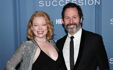 Couple smiling at "Succession" premiere, dressed in formal attire, with blue background displaying logo.