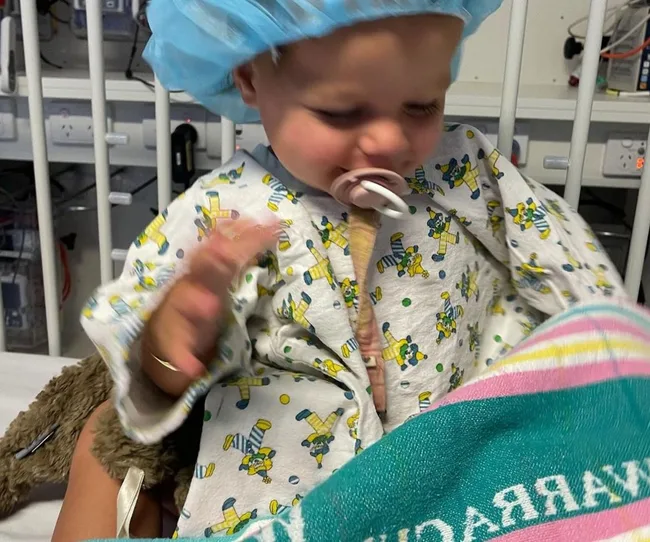 Baby in a hospital gown and surgical cap, sitting with a pacifier in mouth, on what appears to be a hospital bed.