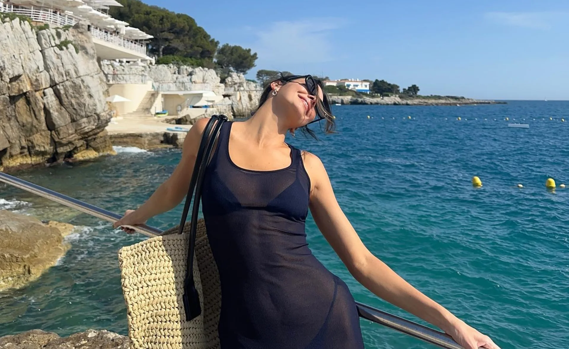Woman in a sheer black dress poses joyfully by the sea with a straw bag on her shoulder.