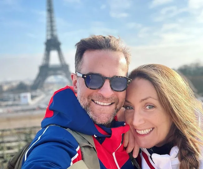 Couple smiling with Eiffel Tower in the background, wearing sunglasses and colorful jackets on a sunny day.