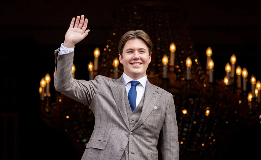Prince Christian of Denmark in a plaid suit waves, smiling, with a chandelier in the background.