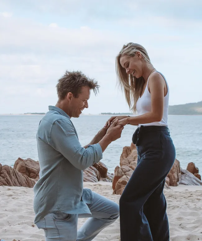A man proposes to a woman on a beach, kneeling with a ring.