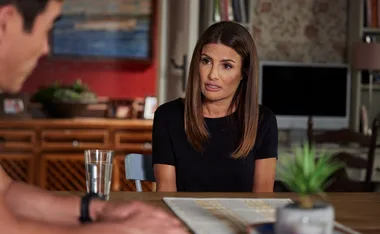 Woman with straight brown hair sitting at a table, engaged in conversation, with a blurred male in the foreground.