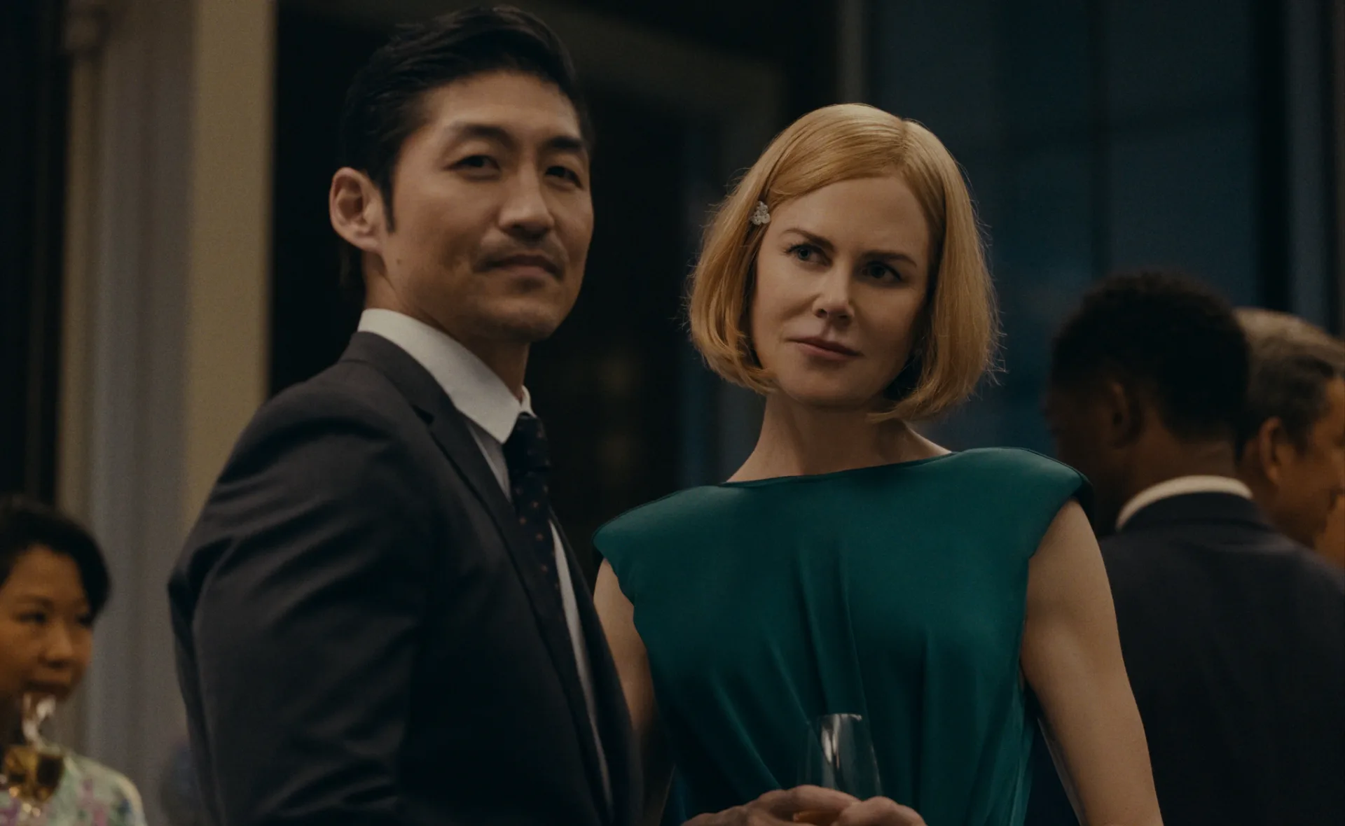 Two people in formal attire at a social event, one holding a drink, with others visible in the background.