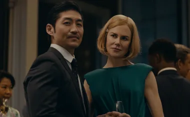 Two people in formal attire at a social event, one holding a drink, with others visible in the background.