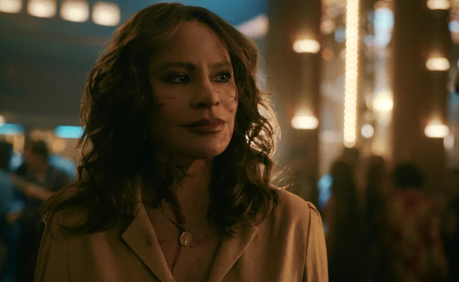 A woman with wavy hair and a necklace in a dimly lit room, faint blood stains visible on her face and clothes.