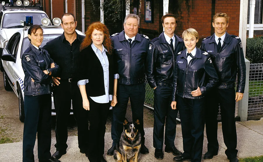 A group of seven officers and a dog standing in front of a police car, wearing uniforms outside a brick building.