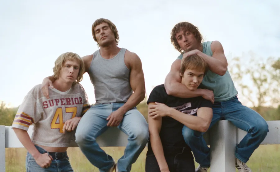 Four young men in casual attire, leaning on a white fence, one wearing a "SUPERIOR 47" jersey, outdoor setting.