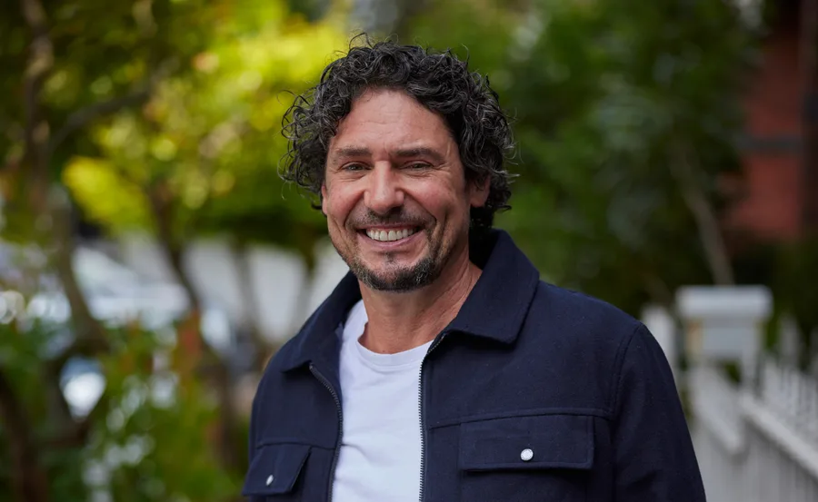 Man with curly hair smiling in a park, wearing a navy jacket over a white shirt.