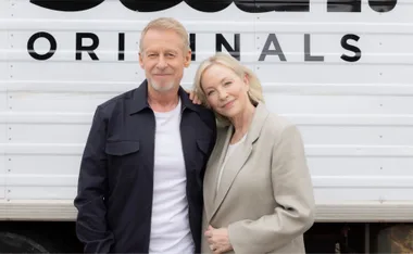 Elderly couple posing in front of a trailer with "ORIGINALS" text behind them, looking content.