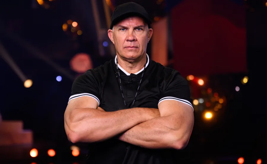 Man in black shirt and cap standing with arms crossed against a blurred background with warm lighting.
