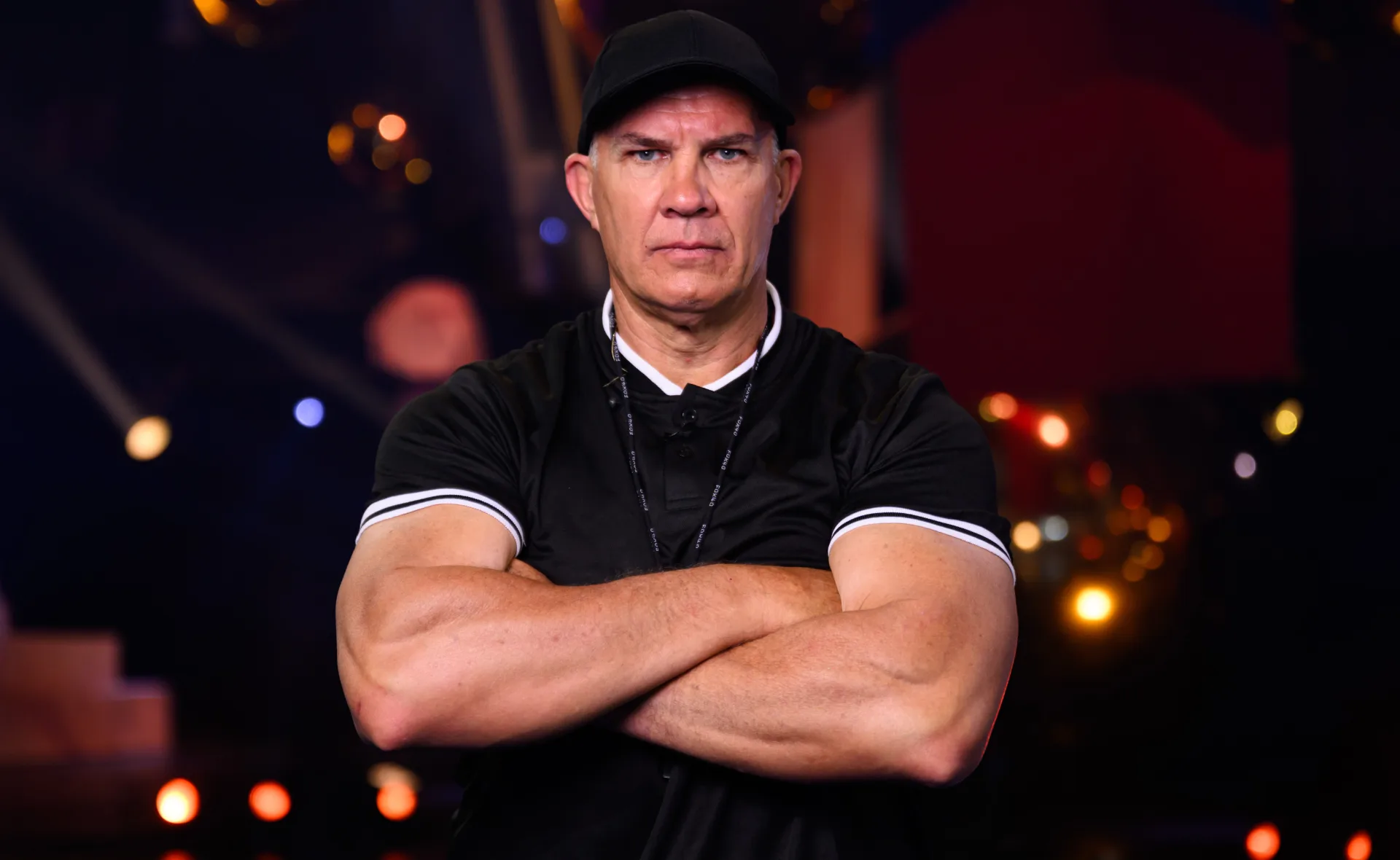 Man in black shirt and cap standing with arms crossed against a blurred background with warm lighting.