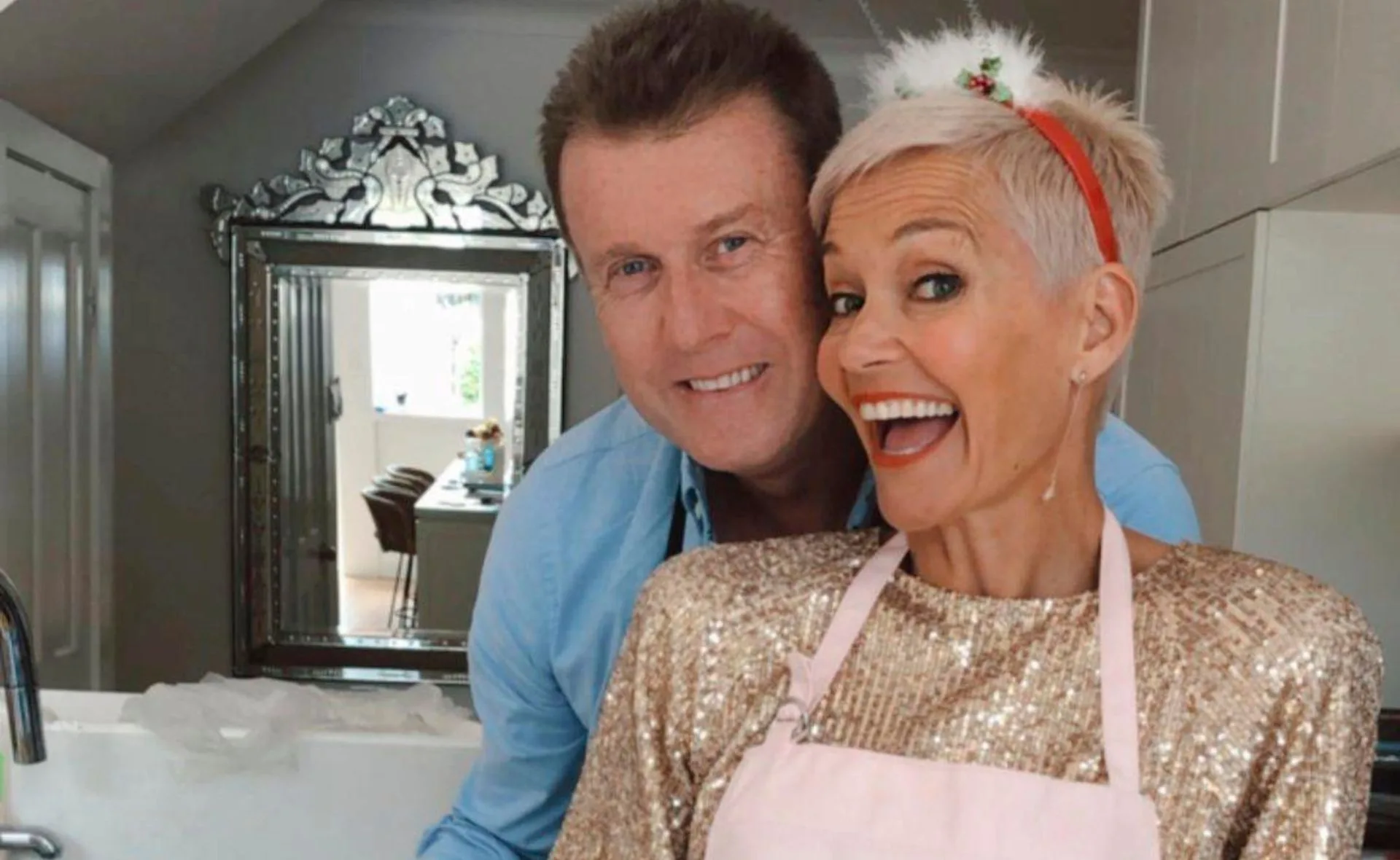 A happy couple in a kitchen, the woman in a shiny gold top and pink apron, smiling widely at the camera.