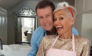 A happy couple in a kitchen, the woman in a shiny gold top and pink apron, smiling widely at the camera.
