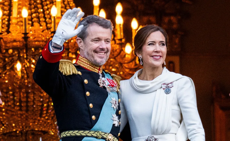 Royal couple in formal attire waving with a chandelier in the background.