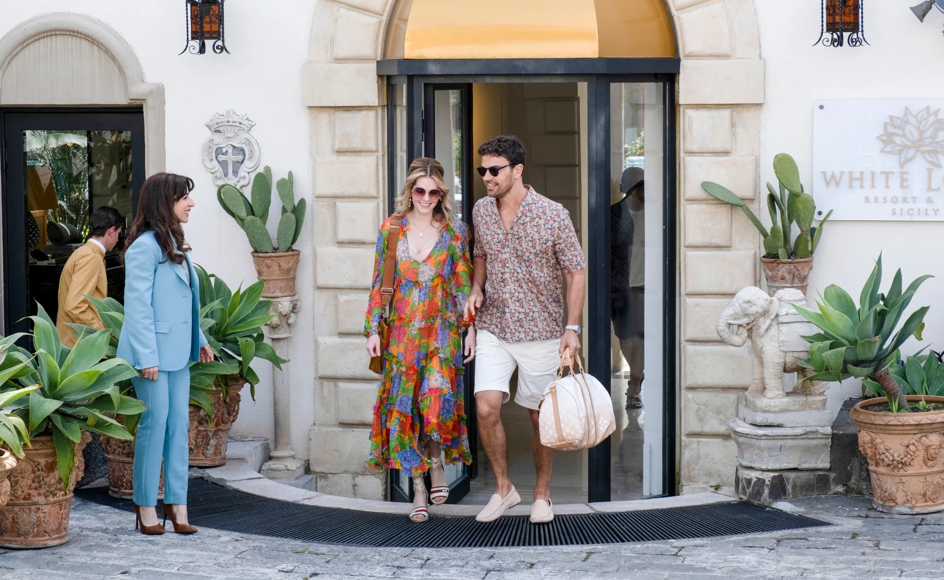 Guests exiting a resort, with a woman in a blue suit, others in summer attire, surrounded by potted plants.