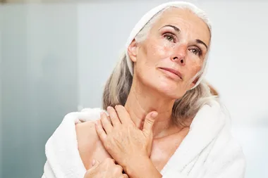 Woman with gray hair and towel touching her neck, looking upwards thoughtfully.