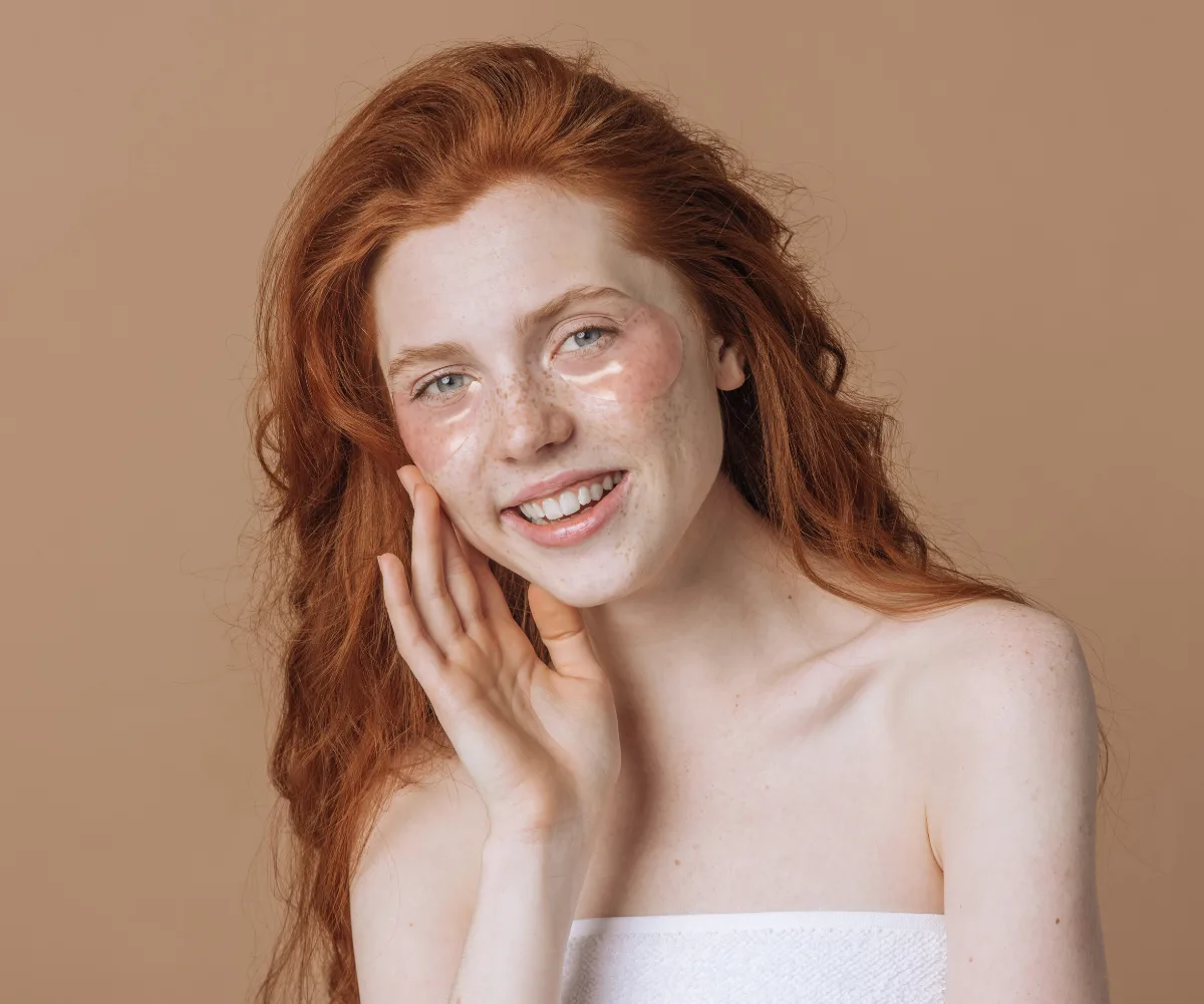 Young woman with long red hair and freckles smiles, wearing under-eye patches, against a beige background.