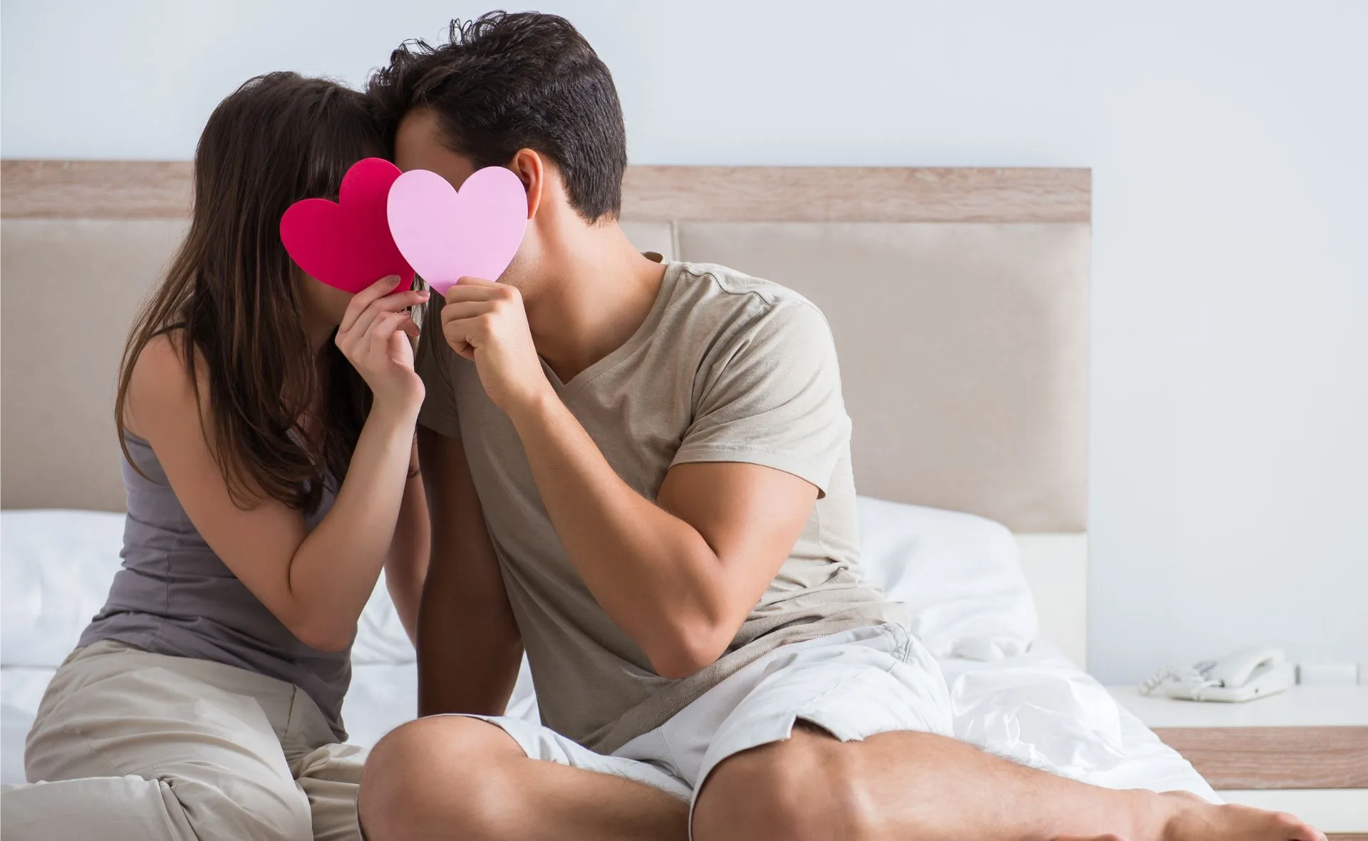 Couple sitting on bed, holding red and pink heart cutouts in front of their faces.