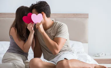 Couple sitting on bed, holding red and pink heart cutouts in front of their faces.