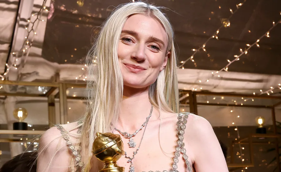 A person with long blonde hair holds a golden globe trophy, smiling against a backdrop of lights.
