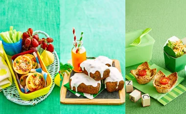 A variety of colorful lunchbox foods including mini quiches, grapes, cupcakes, juice bottle, and salad on a green surface.