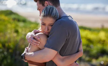 Man and woman hugging emotionally in a sunny outdoor setting by the beach.
