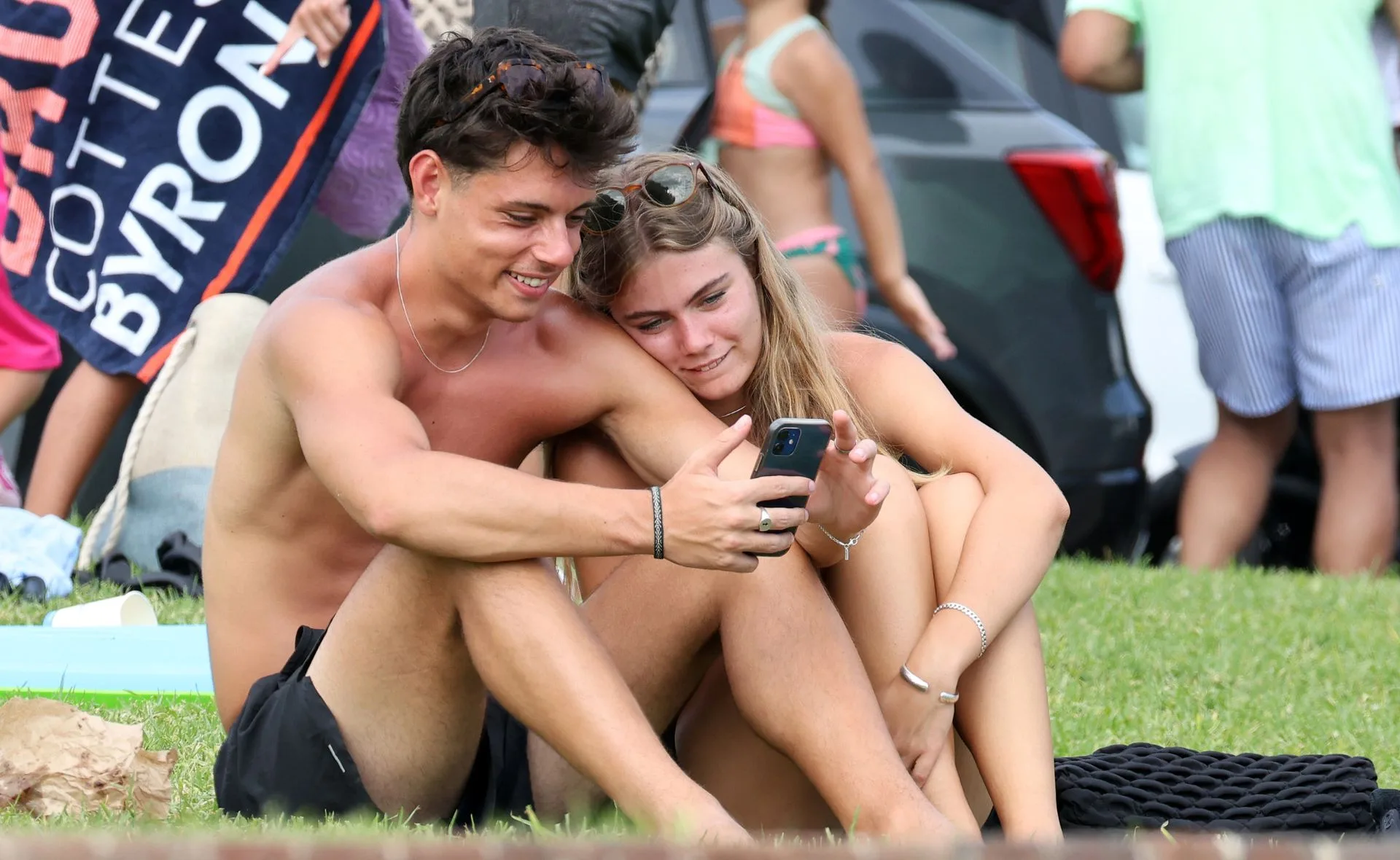 Young couple sitting on grass, smiling and looking at a smartphone together while surrounded by beachgoers.