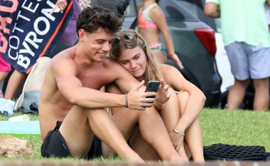 Young couple sitting on grass, smiling and looking at a smartphone together while surrounded by beachgoers.