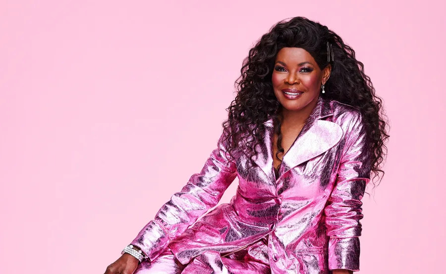 Person in a shiny pink outfit with curly hair, smiling against a pink background.