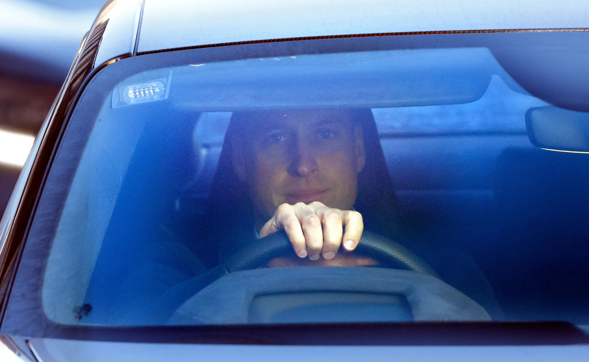 A person driving a car, seen through the windshield, with hands on the steering wheel.