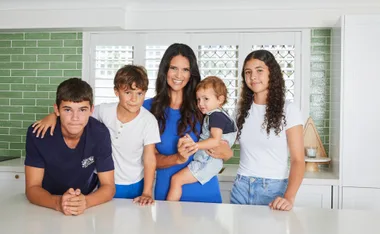 A woman in blue surrounded by four children in a modern kitchen with green tiles.