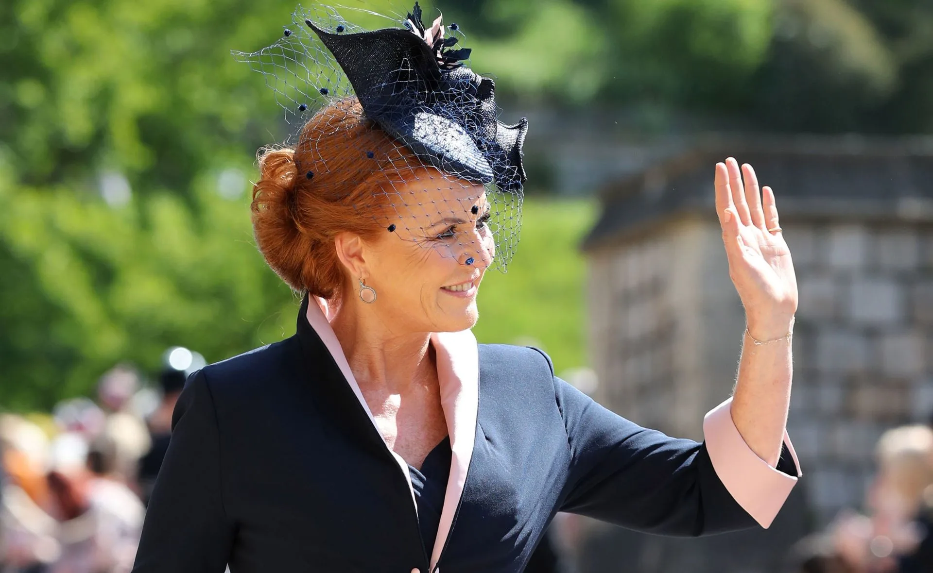 A person with red hair and a black hat waves outdoors, wearing a dark coat with pink lapels.