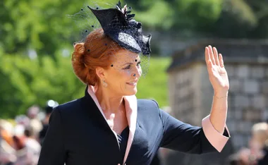 A person with red hair and a black hat waves outdoors, wearing a dark coat with pink lapels.