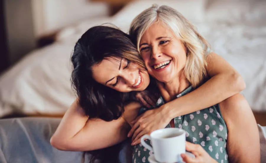 Mother and daughter hugging and smiling, sitting on a couch, with the mother holding a cup of coffee.
