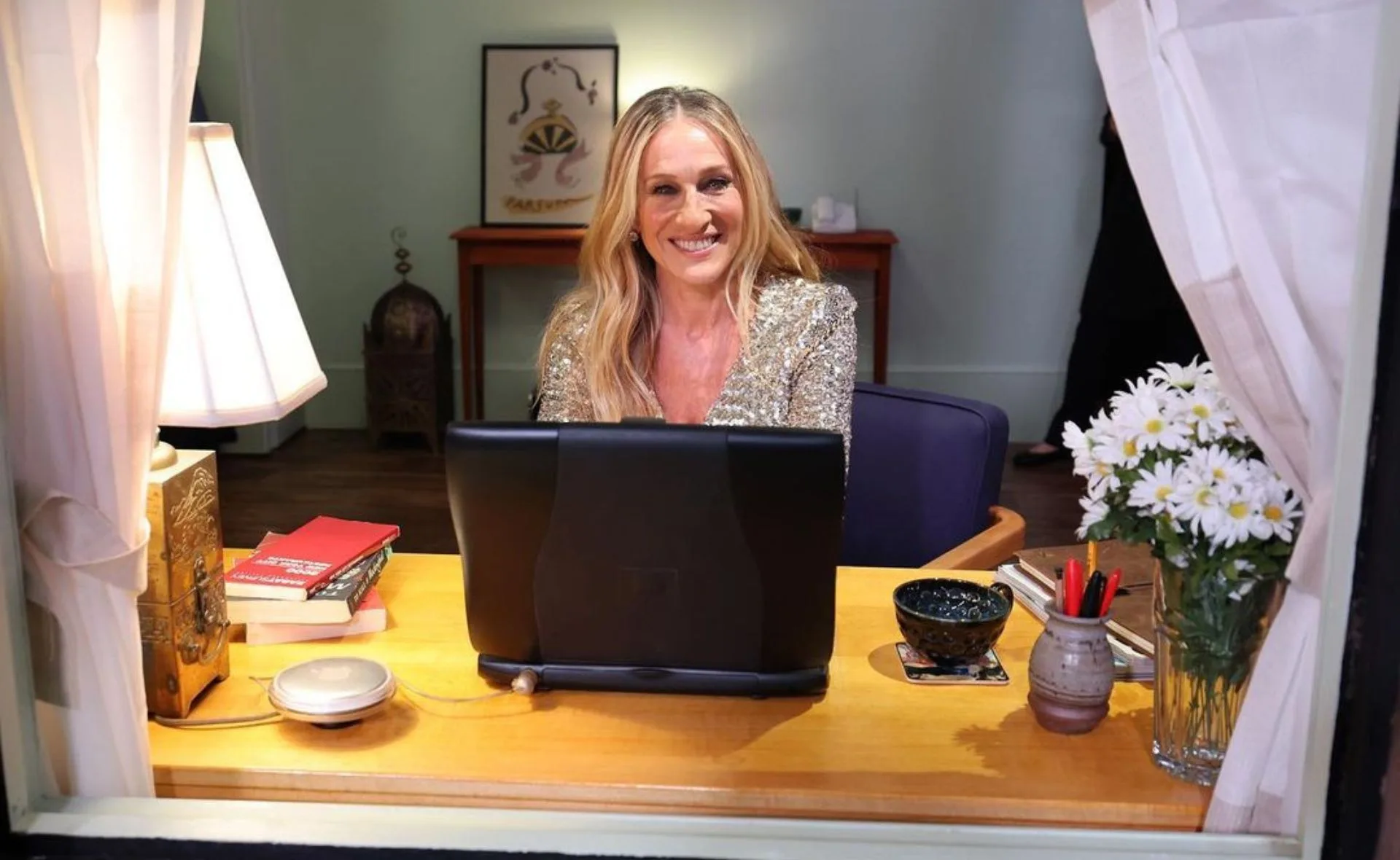 Smiling woman at a desk with a laptop, books, and flowers, viewed through a window.
