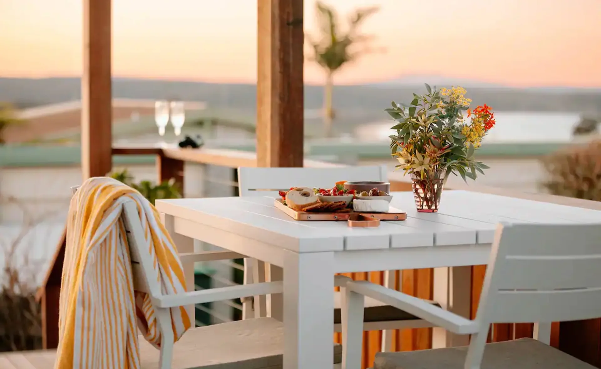 Outdoor dining table with a yellow-striped towel, snacks, and vase of flowers at sunset, overlooking a scenic view.