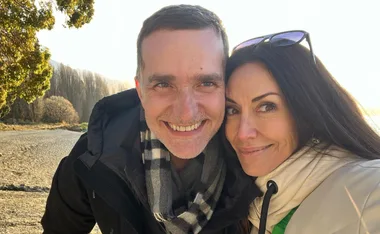 A smiling couple takes a selfie on a sunny beach with trees in the background.