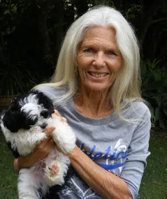 An older woman with long gray hair smiles outdoors, holding a black and white puppy in her arms.