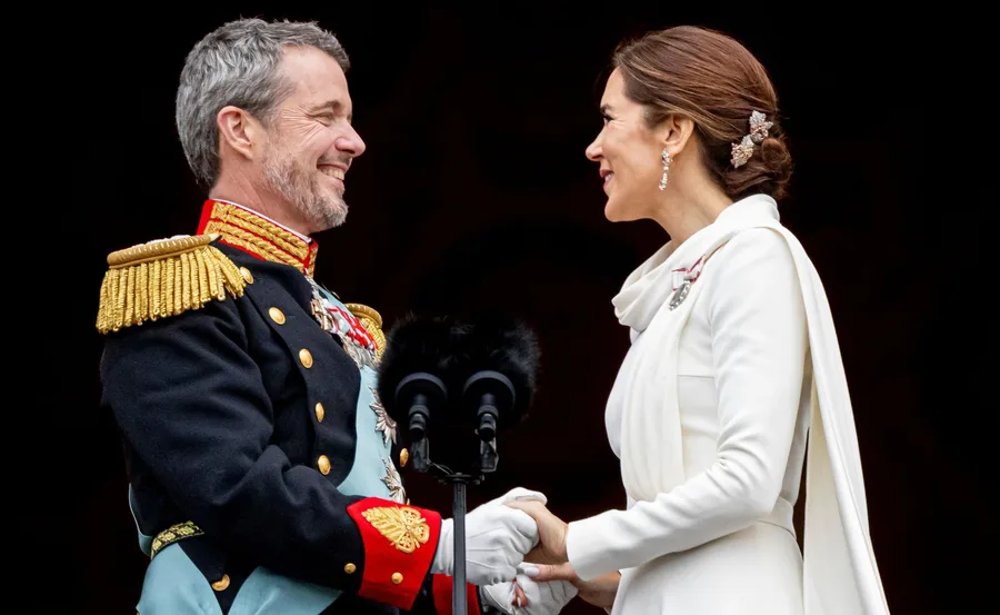 Royal couple smiling and holding hands during an official event, dressed in formal attire.