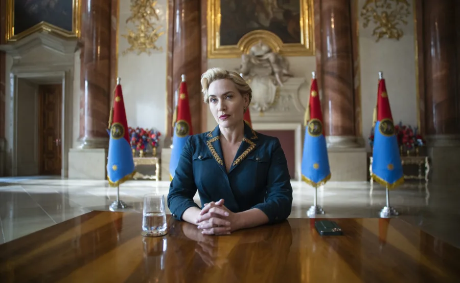 A woman in formal attire sits at a table in an ornate room with flags behind her.