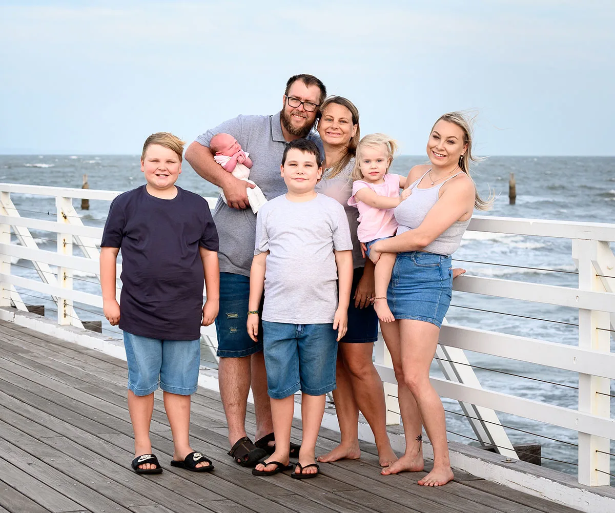 Family of six smiling on a seaside boardwalk, with an ocean view in the background.